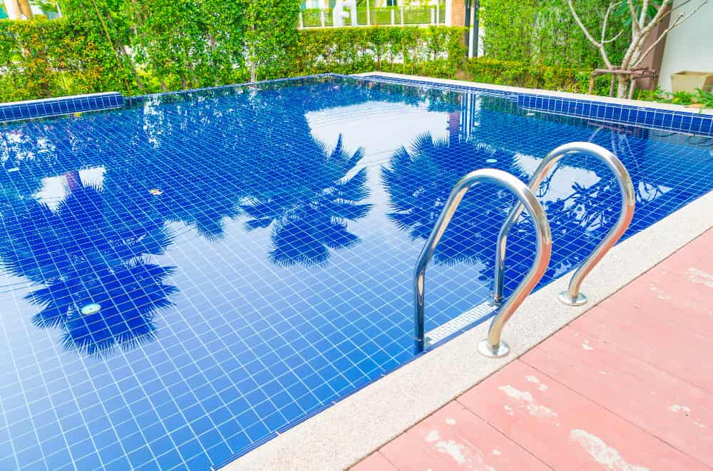 Piscine moderne avec un revêtement en carrelage de piscine bleu foncé, une échelle en inox et des reflets de palmiers sur l'eau.
