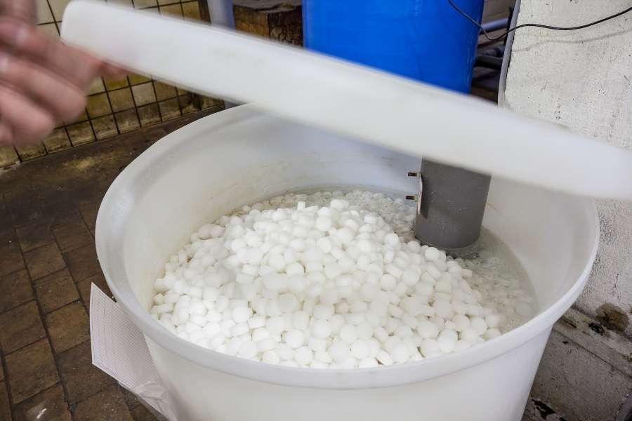 Une main soulève le couvercle d'un bac rempli de pastilles de sel blanches, montrant le réservoir nécessaire pour installer un adoucisseur d'eau efficacement.