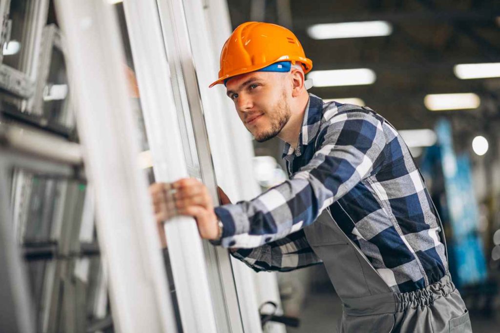 Un ouvrier en casque de chantier inspecte des profilés de fenêtres en PVC dans une usine.